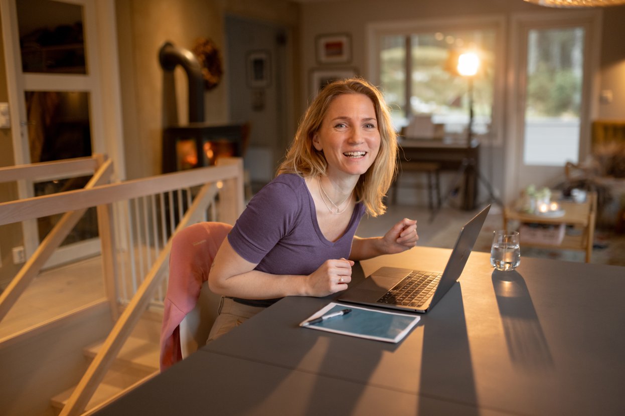 Kvinnelig students sitter ved bordet i stua, jobber på PC og smiler til kameraet